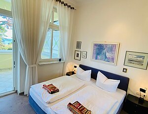 Schlafen mit Meeresrauschen hier im Schlafzimmer der Ferienwohnung 8 in der Villa Caprivi mit blauem Boxspringbett in Komforthöhe, grauem Teppichboden und schwarzen Nachtschränken mit Leselampen. Große Fenster mit bodentiefen, weißen Stores führen zum Balkon Richtung Heringsdorfer Promenade und Ostsee und ermöglichen Seeblick. Über dem Bett hängen dekorative Bilder – freundlich und komfortabel eingerichtet.