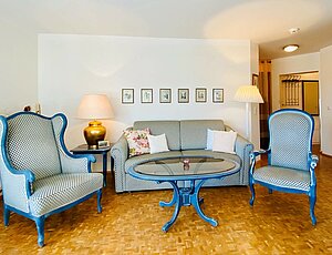 Klassisch-eleganter Wohnbereich der Ferienwohnung 28 in der Residenz Bleichröder mit blauer Couchgarnitur, Holztisch mit Glasplatte und goldenfarben Schirmlampe. Echtholzparkett sorgen für eine warme Atmosphäre.