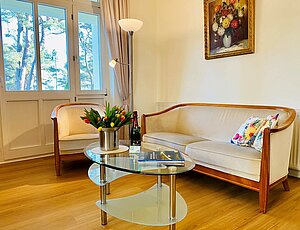 Wohnbereich der Ferienwohnung 3 in der Villa Caprivi mit beigem Samtsofa, zwei passenden Sesseln und modernem Glastisch mit Blumenvase. Stehlampe neben der Sitzgruppe, pflegeleichter Boden in Holzoptik. Große Fensterfront mit Tür zum überdachten Balkon Richtung Ostsee – klassisch eingerichtet, hell und einladend.
