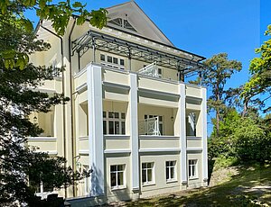 Repräsentative Bäderstilvilla Caprivi an der Heringsdorfer Promenade mit Ferienwohnungen, die vom Vermietungsservice Sass vermarktet werden. Freistehende Villa Caprivi mit großzügigen Fenstern, kannelierten Säulen, überdachten Balkonen, in einem hellen Cremeton gestrichen. 