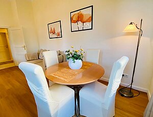 Runder Esstisch mit hölzerner Platte in der Ferienwohnung 3 der Villa Caprivi, dekoriert mit weißer Vase und Kunstblumen. Vier Stühle mit weißen Hussen, eine Stehlampe sorgt für zusätzliches Licht. An der Wand hängen große Bilder mit Blumenmotiven – helles, harmonisches Ambiente im Wohnbereich.