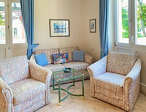 Gemütliche Sitzecke in der Ferienwohnung 10 der Villa Caprivi mit beiger Zweisitzercouch und zwei Sesseln im floralen Muster, blauen Dekokissen, rechteckigem Glastisch sowie großen Fenstern mit bodenlangen blauen Gardinen – heller, harmonisch gefliester Wohnbereich in klassischem Stil.