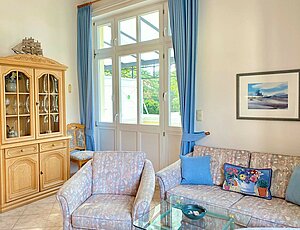 Willkommen in der Ferienwohnung 10 der Villa Caprivi mit  gemütlichem Wohnbereich, großer Fensterfront und bodentiefen, blauen Vorhängen. Neben dem Fenster stehen eine gemusterte Couch und ein passender Sessel mit blauen Dekokissen. In der Mitte befindet sich ein Glastisch. Links ergänzt ein heller Holzvitrinenschrank mit Geschirr den Raum. Durch die Tür ist der Balkon mit Blick ins Grüne sichtbar – freundlich und klassisch eingerichtet.