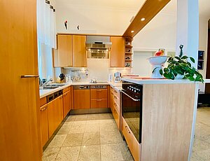 Moderne U-förmige Komplettküche in der Ferienwohnung 8 der Villa Caprivi mit hellen Buchenholzfronten und Edelstahlgriffen, integriert in den Wohnraum. Ausgestattet mit Geschirrspüler, Waschmaschine, Backofen, Ceranfeld mit Dunstabzugshaube, Kaffeemaschine und Wasserkocher. Ein Tresen nutzbar als Esstisch, dekoriert mit Grünpflanze und Obstschale. Eine weiße Säule mit beleuchtetem Baldachin bildet den stilvollen Abschluss.