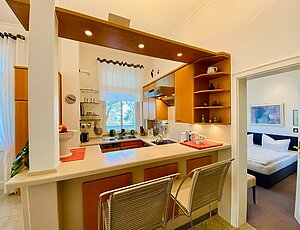 Ferienwohnung 8 Villa Caprivi direkt an der Promenade Heringsdorfs mit seiner offenen Küche. Diese ist U-förmig angelegt und komplett ausgestattet. Auf dem Tresen stehen eine Obstschale und eine Glasschale, davor zwei Metallbarhocker. Links bildet eine weiße Säule den Küchenabschluss, nach oben ein beleuchteter Baldachin. Durch ein Fenster ist die Heringsdorfer Promenade und durch die alten Kiefern das Meer zu sehen. Rechts führt eine Tür ins Schlafzimmer mit Doppelbett. 