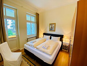 Schlafzimmer der Ferienwohnung 3 in der Villa Caprivi mit großem Doppelbett in Komforthöhe auf Echtholzparkett, weiß bezogen und mit bereitliegenden Handtüchern. Zugang zur überdachten Terrasse mit Blick auf die Promenade 