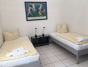 Helles Schlafzimmer der Ferienwohnung 5 der Villa Caprivi mit zwei flexibel zusammenschiebbaren grauen Einzelbetten, schwarzen Edelstahl-Nachttischen mit Glasplatte und Lampen, gerahmtem Kunstplakat über den Betten sowie weiß gefliestem Boden.
