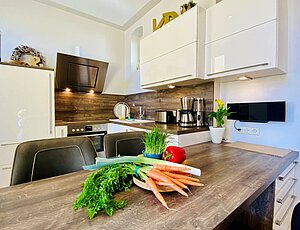 Moderne, offen gestaltete Küche in der Ferienwohnung Kaiserperle mit weißen Hochglanzschränken, Ceranfeld, Backofen, Geschirrspüler, Kaffeemaschine und Toaster. Angrenzender Essbereich mit vier Lederstühlen, Gemüseschale auf dem Tisch, Musikanlage an der Wand – stilvoll und komplett ausgestattet für den Urlaub.
