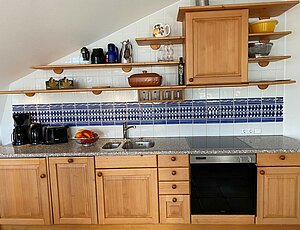 Gemütliche Echtholzküche von Domizil im Wohnzimmer der Ferienwohnung 12, ausgestattet mit Backofen, Ceranfeld, Geschirrspüler und Kaffeemaschine; weiße Fliesen und eine blaue Bordüre, offene Regale, harmonisch in die Dachschräge integriert.