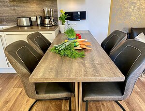 Esstisch der Ferienwohnung Kaiserperle im Übergang zwischen Küche und Wohnbereich, mit hölzerner Tischplatte auf Metallbeinen, vier bequemen Ledersesseln und einer Schale frischem Gemüse. Musikanlage an der Wand sowie Kaffeemaschine und Toaster sind vom Tisch aus leicht erreichbar – modernes, funktionales Design für entspannten Urlaub.
