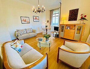 Wohnzimmer der beliebten Ferienwohnung 3 in der Villa Caprivi, eingerichtet in Beige und heller Holzoptik. Sitzgruppe aus beiger Samtcouch, Clubsessel und Glastisch, dahinter eine zusätzliche Schlafcouch. Gegenüber steht eine halbhohe Kommode mit Glastüren und Schubladen, daneben ein TV auf einem kleinen Schränkchen. Über der Kommode hängt ein dunkles Bild. Im Hintergrund ein runder Esstisch mit vier Stühlen vor der Glastür zum zweiten Balkon. Ein Kronleuchter aus Messing hängt zentral von der Decke.