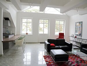 Moderne Ferienwohnung 11 im Dachgeschoss der Villa Caprivi mit freiliegenden weißen Dachbalken, großer dreiteiliger Fensterfront mit Blick ins Grüne und Richtung See, schwarzen Ledercouchen auf rot gemustertem Wollteppich, Glastisch sowie offenem Küchenbereich mit Tresen und vier Schalenstühlen – lichtdurchfluteter, großzügiger Wohnraum.