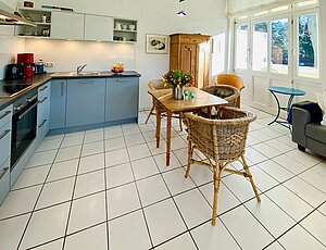 Wohnzimmer der Ferienwohnung 5 in der Villa Caprivi mit moderner graublauer Küchenzeile an der linken Wand, ausgestattet mit Backofen, Cerankochfeld und Dunstabzugshaube, Geschirrspüler und separatem Kühlschrank. Im Zentrum steht ein hölzerner Esstisch mit drei Rattanstühlen, im Hintergrund ein Holzschrank – stilvoller Kontrast zur weißen Einrichtung.