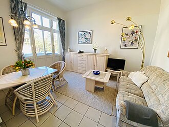 Willkommen in der Ferienwohnung 7der Villa Caprivi mit freundlichem Wohnbereich. großer Esstisch mit Rattanstühlen, bequeme Couch mit Couchtisch, TV auf Kommode und Stehlampe.